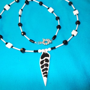 May include: A necklace featuring a white and black patterned pendant shaped like a leaf or shell. The chain consists of alternating white square beads, small black cylindrical beads, and faceted black round beads, finished with a silver toggle clasp.