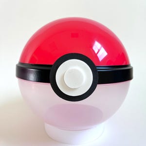 May include: A red and white Poke Ball container with a black band around the middle. The container is made of clear plastic and has a white base.