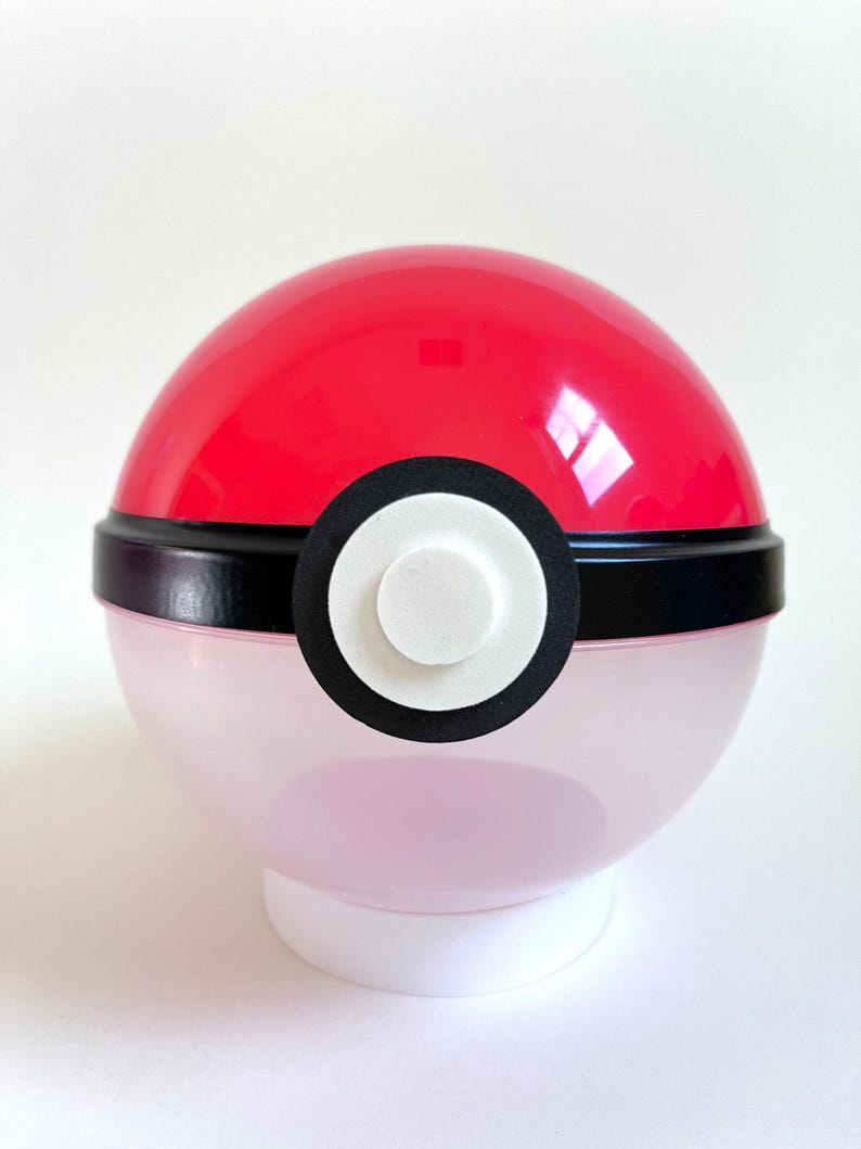 May include: A red and white Poke Ball container with a black band around the middle. The container is made of clear plastic and has a white base.