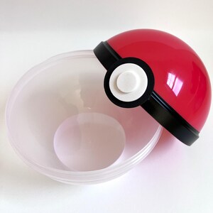 May include: A red and black Poke Ball container with a clear plastic bowl. The Poke Ball has a white button on the front.