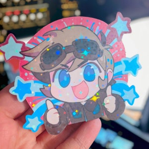 May include: A holographic sticker featuring a cartoon character with brown hair, blue eyes, and a happy expression. The character is wearing goggles and a brown jacket. The sticker is surrounded by blue and pink stars and streaks of light.