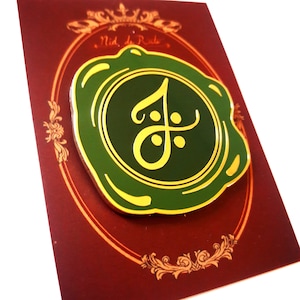 May include: A gold-colored enamel pin with a green wax seal design. The seal features a stylized letter "J" in gold with a small dot on the top and bottom. The text "Nid de Role" is visible in the background.