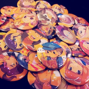 May include: A collection of colorful round buttons featuring various cartoon characters and designs. The buttons are made of metal and have a shiny finish.