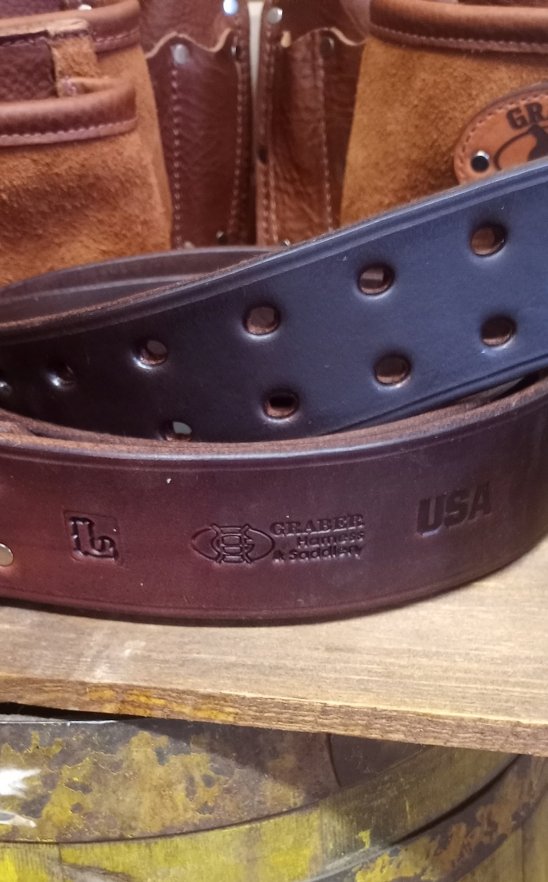 K&ouml;nnte beinhalten: Ein brauner Lederg&uuml;rtel mit einem schwarzen Lederg&uuml;rtel dar&uuml;ber. Der G&uuml;rtel hat eine Messingschnalle und der Text "Graber Harness & Saddlery USA" ist in das Leder gepr&auml;gt.