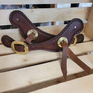 Pu&ograve; includere: Due cinturini per speroni in pelle marrone con fibbie e punte in metallo dorato. I cinturini sono su una superficie di legno.