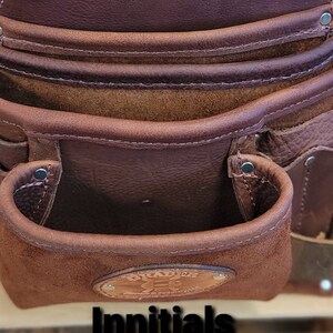 May include: Close-up of a brown leather tool belt with embossed initials "AJR" at the top. The belt has multiple pockets and a suede-lined pouch. The text "Innitials" and color options are visible.
