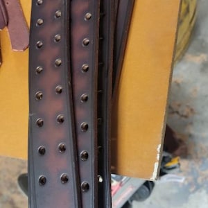 May include: Close-up of multiple brown leather straps with rows of evenly spaced holes. The straps are stacked on top of each other and have a slightly rounded edge.