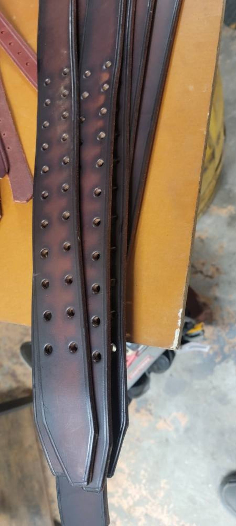 May include: Close-up of multiple brown leather straps with rows of evenly spaced holes. The straps are stacked on top of each other and have a slightly rounded edge.