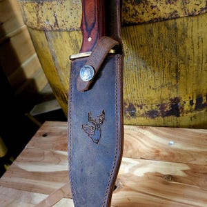 May include: A large hunting knife with a dark wood handle and a leather sheath. The sheath is brown with a deer head design and a metal clasp. The knife is displayed on a wooden surface.
