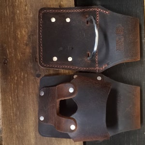K&ouml;nnte beinhalten: Zwei braune Leder-Werkzeugg&uuml;rtel mit Metallnieten und einer Metallschlaufe. Der G&uuml;rtel links hat rote N&auml;hte und den Text "The Tool Belt" aufgedruckt. Der G&uuml;rtel rechts hat eine einzelne Schlaufe auf der Vorderseite.