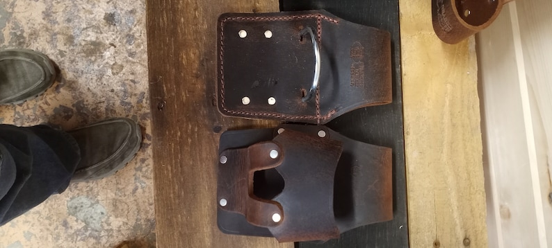K&ouml;nnte beinhalten: Zwei braune Leder-Werkzeugg&uuml;rtel mit Metallnieten und einer Metallschlaufe. Der G&uuml;rtel links hat rote N&auml;hte und den Text "The Tool Belt" aufgedruckt. Der G&uuml;rtel rechts hat eine einzelne Schlaufe auf der Vorderseite.