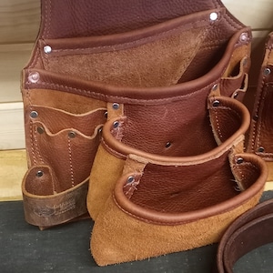 K&ouml;nnte beinhalten: Ein brauner Leder-Werkzeugg&uuml;rtel mit mehreren Taschen und einer Hammerschlaufe. Der G&uuml;rtel ist aus hochwertigem Leder gefertigt und hat eine robuste Konstruktion.