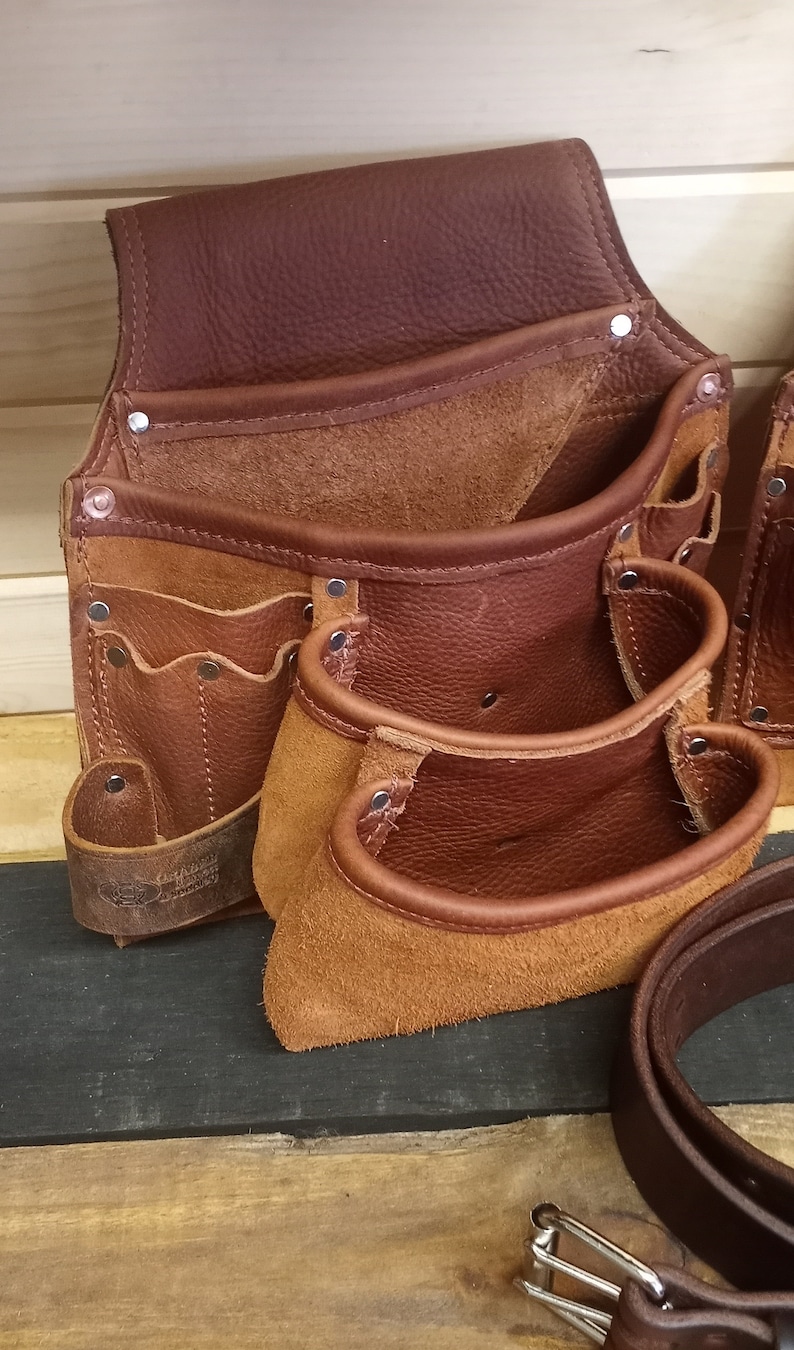 K&ouml;nnte beinhalten: Ein brauner Leder-Werkzeugg&uuml;rtel mit mehreren Taschen und einer Hammerschlaufe. Der G&uuml;rtel ist aus hochwertigem Leder gefertigt und hat eine robuste Konstruktion.