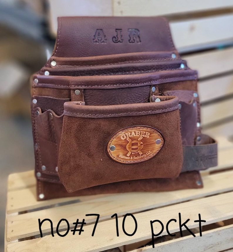 May include: A brown leather tool belt with multiple pockets and rivets. The belt has the initials "AJR" embossed on the top flap and a "GRABER" logo on the front pocket. The text "no#7 10 pckt" is written at the bottom.