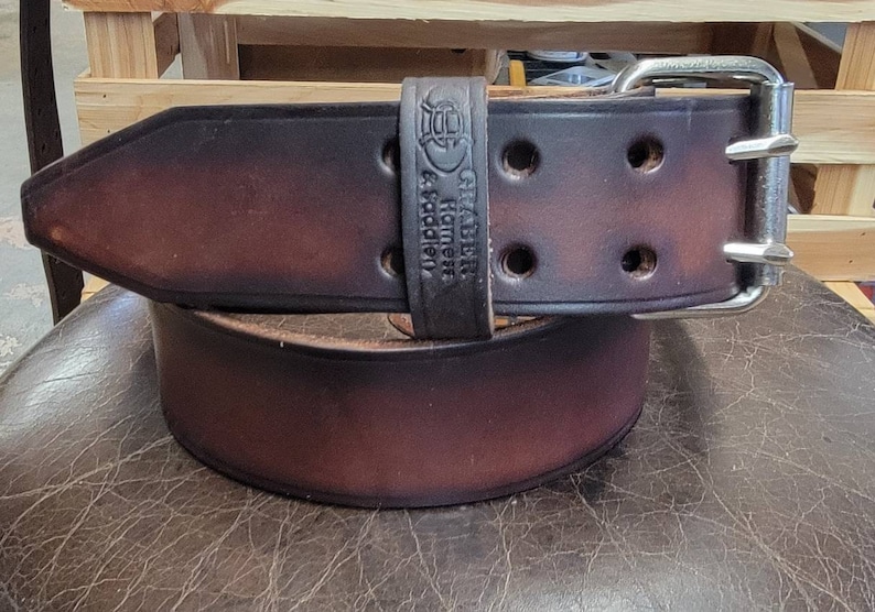 May include: A brown leather belt with a silver buckle. The belt has a distressed look and is embossed with the text "GRABBER Harness & Saddlery".