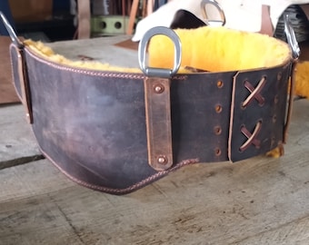 May include: A brown leather work belt with a yellow fleece lining and two metal D-rings. The belt has a stitched design and is secured with a buckle.