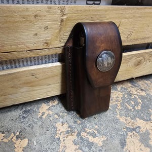 May include: A brown leather case with a flap closure and a silver buffalo nickel detail. The case is rectangular with a stitched edge and appears to be handmade. The background includes a wooden crate and newspaper clippings.