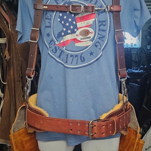 May include: A brown leather tool belt with a brown leather strap and buckle. The belt has two brown leather pouches on the sides. The belt is worn over a blue t-shirt with the text "FREEDOM RING EST. 1776" and an American flag graphic.