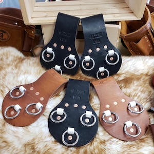 Puede incluir: Varios portadores de herramientas de cuero en tonos marrones y negros, cada uno con anillos y remaches metálicos. Los soportes están estampados con "USA" y un logotipo.