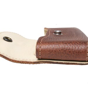 May include: A brown leather case with a flap that opens to reveal a white interior. The case has a metal snap closure.