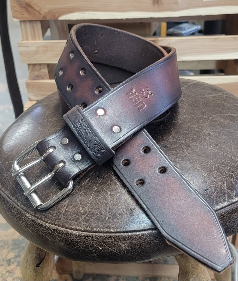 May include: A brown leather belt with a silver buckle and double rows of stitching. The belt has a distressed look and the text "S. Van" is embossed on the leather.