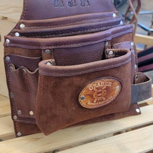 May include: A brown leather tool belt with multiple pockets and a suede pouch. The top flap is embossed with "AJR". A wooden oval patch on the front pouch displays the "GRABER" logo. Rivets secure the leather pieces.