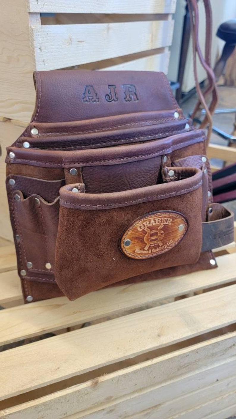 May include: A brown leather tool belt with multiple pockets and a suede pouch. The top flap is embossed with "AJR". A wooden oval patch on the front pouch displays the "GRABER" logo. Rivets secure the leather pieces.