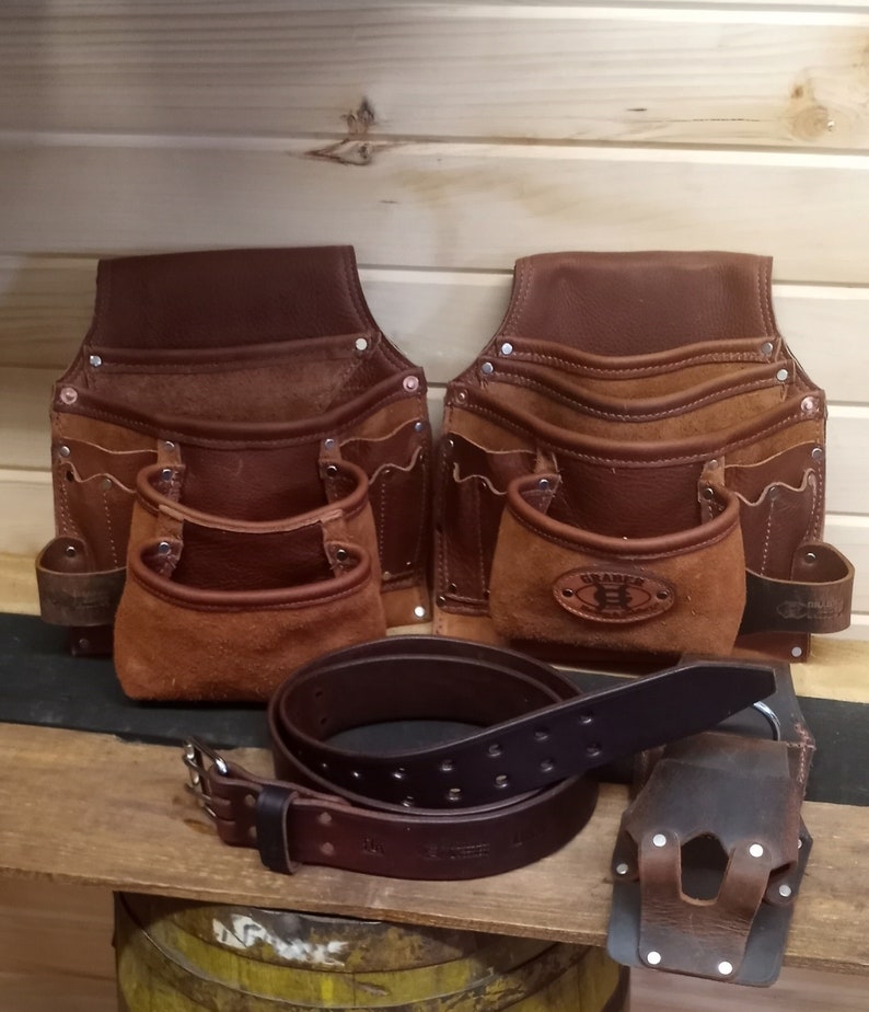 K&ouml;nnte beinhalten: Zwei braune Leder-Werkzeugg&uuml;rtel mit mehreren Taschen und ein brauner Lederg&uuml;rtel mit einer silbernen Schnalle. Die Werkzeugg&uuml;rtel sind mit Wildleder gef&uuml;ttert und der G&uuml;rtel ist aus doppeltem Leder.
