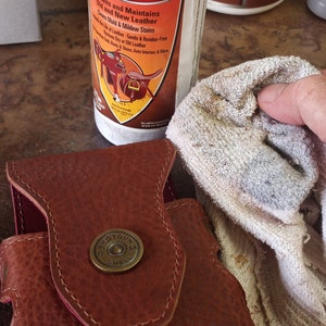 May include: A white bottle of Leather Therapy Restorer & Conditioner with a brown label featuring a saddle and a bull. The bottle is being used to clean a brown leather holster with a brass shotgun shell button.