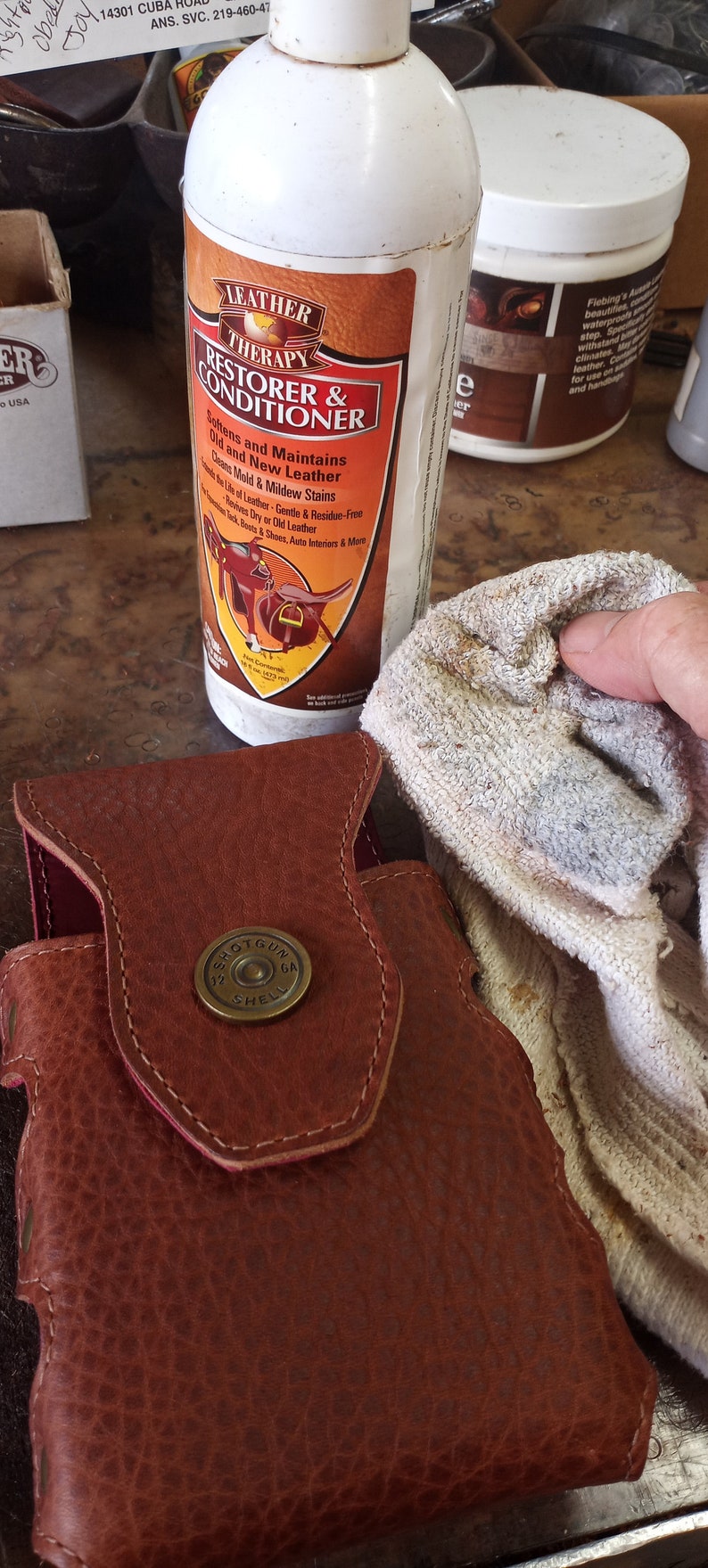 May include: A white bottle of Leather Therapy Restorer & Conditioner with a brown label featuring a saddle and a bull. The bottle is being used to clean a brown leather holster with a brass shotgun shell button.