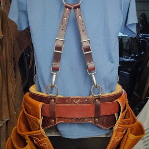 K&ouml;nnte beinhalten: Ein brauner Leder-Werkzeugg&uuml;rtel mit mehreren Taschen und einem passenden braunen Lederschellen-System. Der G&uuml;rtel hat einen braunen Lederriemen mit einer silbernen Schnalle. Die Hosentr&auml;ger haben einen silbernen Ring oben und silberne Clips unten.