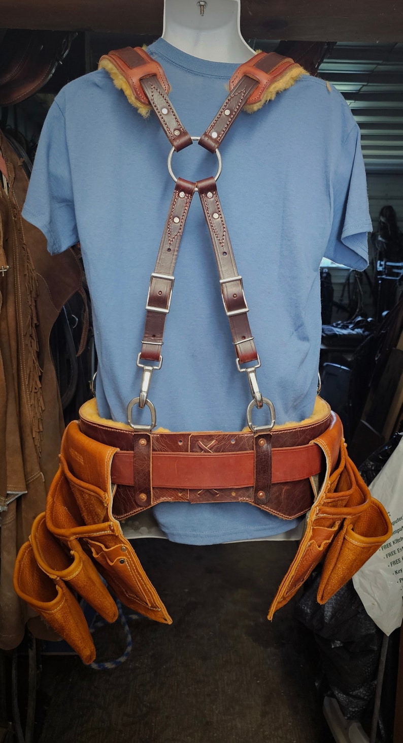 K&ouml;nnte beinhalten: Ein brauner Leder-Werkzeugg&uuml;rtel mit mehreren Taschen und einem passenden braunen Lederschellen-System. Der G&uuml;rtel hat einen braunen Lederriemen mit einer silbernen Schnalle. Die Hosentr&auml;ger haben einen silbernen Ring oben und silberne Clips unten.