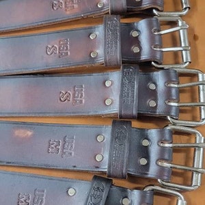 May include: Six brown leather belts with silver buckles. The belts have the text "USA" and "S" embossed on them. The belts are arranged in a stack, with the buckles facing the viewer.