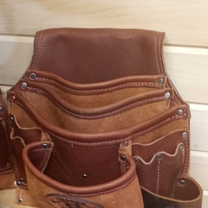 K&ouml;nnte beinhalten: Braunes Leder-Werkzeugg&uuml;rtel mit mehreren Taschen und einem Wildlederboden. Der G&uuml;rtel hat einen braunen Lederriemen mit dem Text "Gelar Leather" darauf.