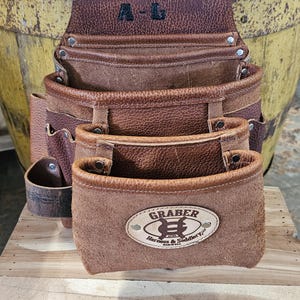 May include: Brown leather tool belt with multiple pockets and a Graber Harness & Saddlery Co. logo. The belt has a personalized inscription "A-L".