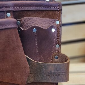 May include: Close-up of a brown leather tool belt with multiple pockets and rivets. The belt features a strap with a logo and the text "Barns & Saddlery". The leather has a distressed look.