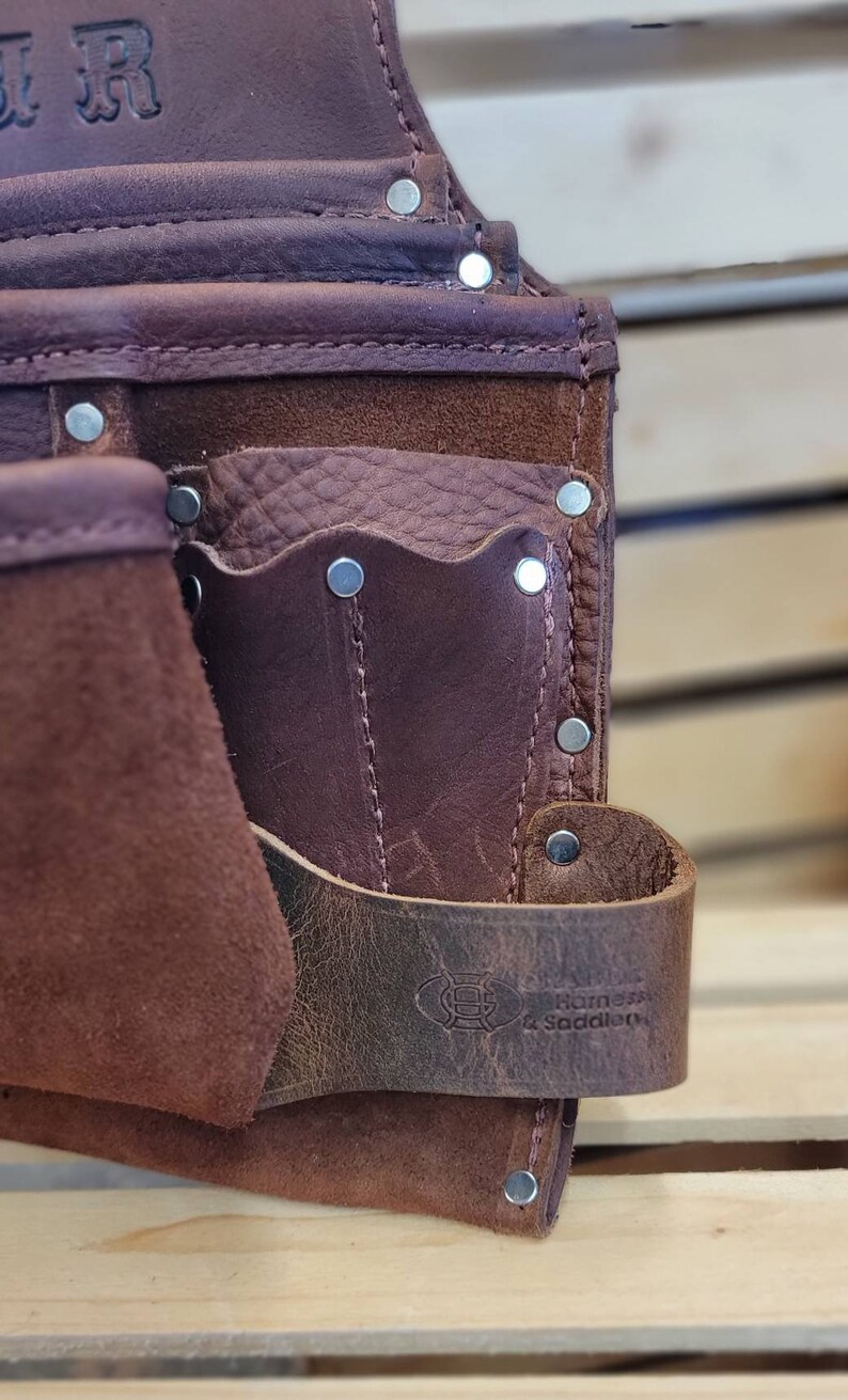 May include: Close-up of a brown leather tool belt with multiple pockets and rivets. The belt features a strap with a logo and the text "Barns & Saddlery". The leather has a distressed look.
