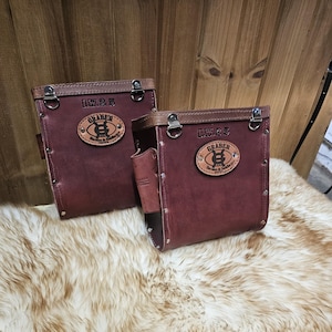 May include: Two burgundy leather tool pouches with brown trim and silver hardware. Each pouch features a leather patch with the text "I.W. 25" and "GRABER Harness & Saddlery". The pouches are on a fluffy, light-coloured surface.