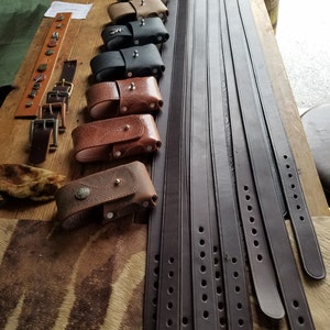 May include: A collection of brown leather belts with multiple rows of stitching and holes. The belts are arranged in a fan shape on a wooden surface. There are also several small leather pouches with metal accents.