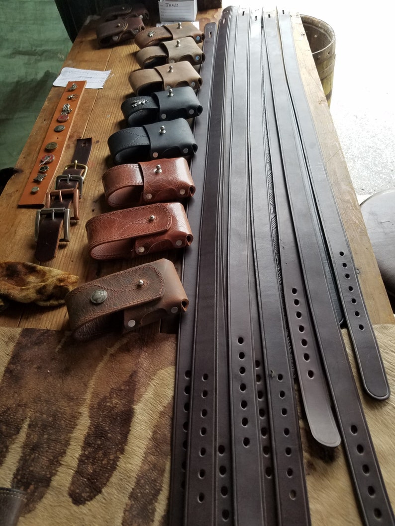 May include: A collection of brown leather belts with multiple rows of stitching and holes. The belts are arranged in a fan shape on a wooden surface. There are also several small leather pouches with metal accents.