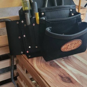 May include: Black leather tool belt with multiple pockets and loops. The belt has a brown leather patch with the text "Glazer" and a graphic of a hammer and a wrench. The belt is sitting on a wooden surface.