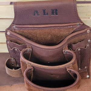 K&ouml;nnte beinhalten: Braunes Leder-Werkzeugg&uuml;rtel mit mehreren Taschen und Schlaufen. Der G&uuml;rtel hat die Initialen "ALR" auf der Vorderseite gepr&auml;gt.