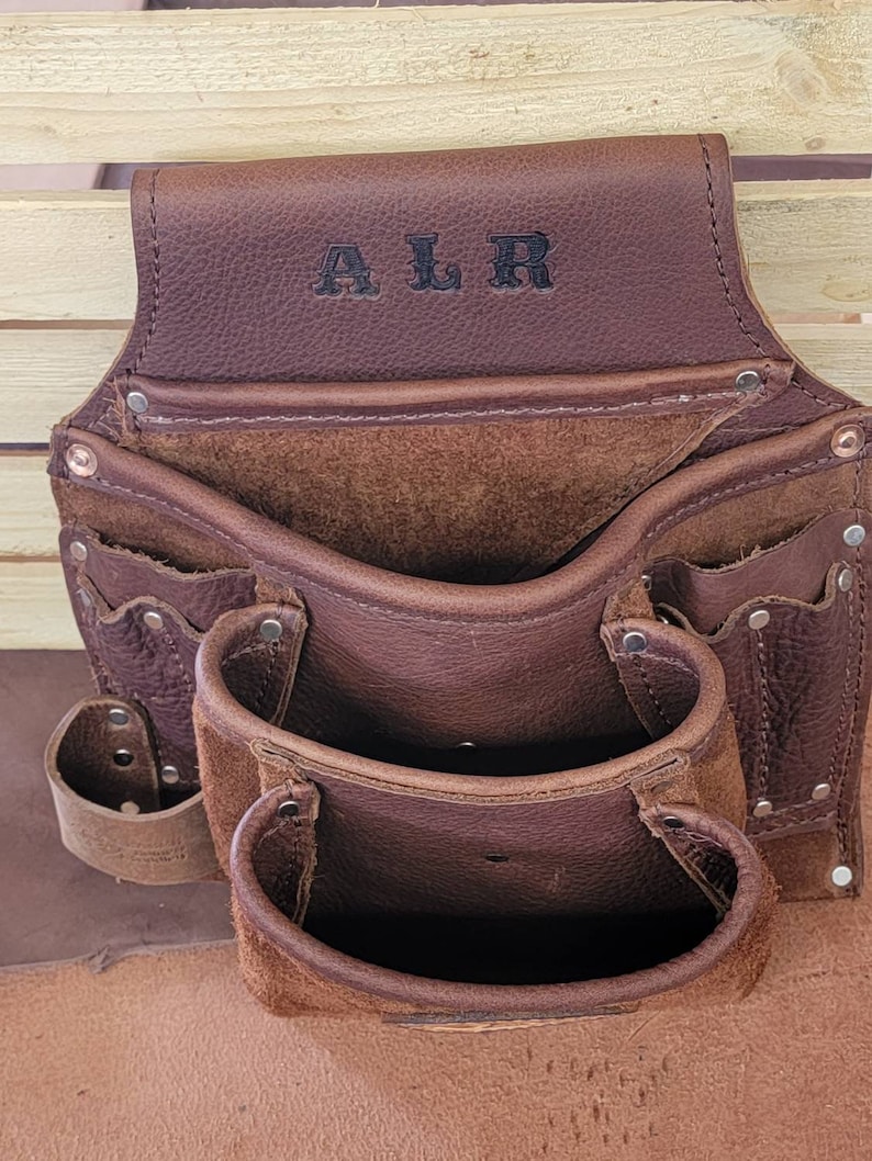 K&ouml;nnte beinhalten: Braunes Leder-Werkzeugg&uuml;rtel mit mehreren Taschen und Schlaufen. Der G&uuml;rtel hat die Initialen "ALR" auf der Vorderseite gepr&auml;gt.