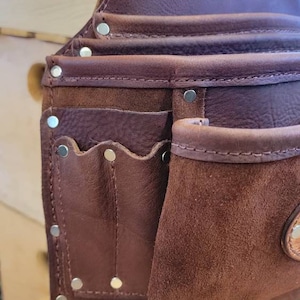 May include: Close-up of a brown leather tool belt with multiple pockets and rivets. The belt has a suede pocket and the initials "AJR" embossed on the top. The leather has a rich, textured appearance, suggesting durability and quality.