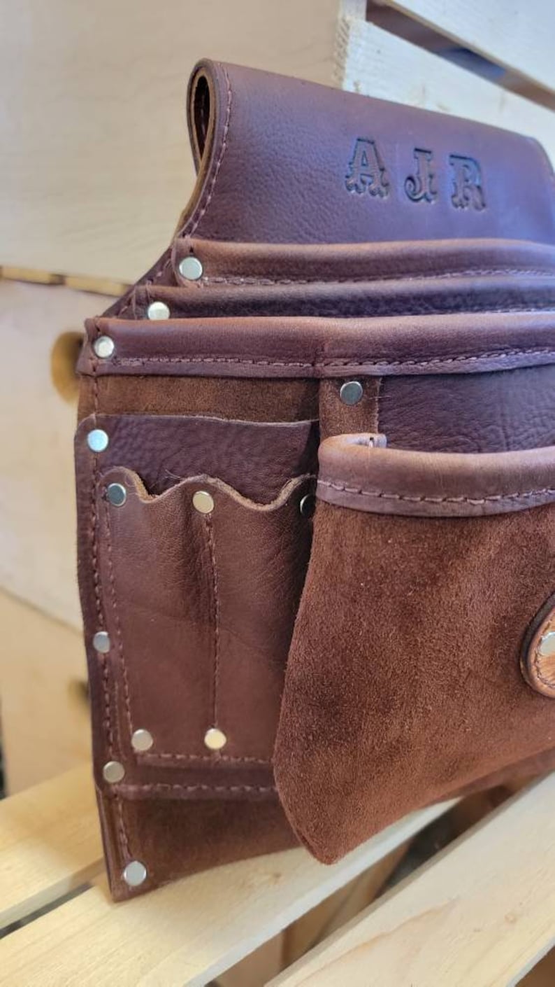 May include: Close-up of a brown leather tool belt with multiple pockets and rivets. The belt has a suede pocket and the initials "AJR" embossed on the top. The leather has a rich, textured appearance, suggesting durability and quality.