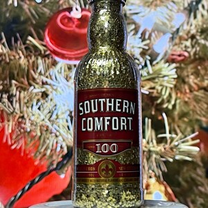 May include: A gold glitter-filled bottle ornament shaped like a miniature bottle of Southern Comfort 100 proof liquor. The bottle has a red label with gold lettering and a red ribbon.