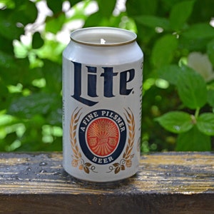 May include: A white Miller Lite beer can candle with navy blue lettering and a gold and navy blue emblem. The candle is set against a backdrop of green foliage and a wooden surface.