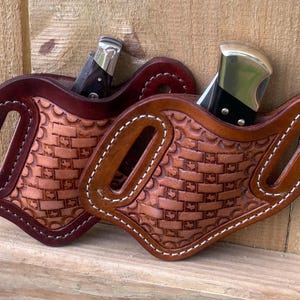 May include: Two leather knife sheaths with a basket weave pattern. One is a deep burgundy colour, and the other is a medium brown. Both have a knife partially inserted. The sheaths have a loop for a belt.