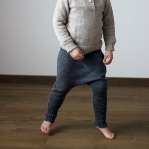 Könnte beinhalten: Ein kleines Kind trägt einen hellbraunen Pullover mit Holzknöpfen und eine dunkelgraue Strickhose. Das Kind lächelt und schaut in die Kamera.