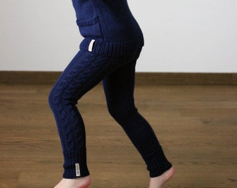 Puede incluir: Una niña pequeña con un suéter azul marino y leggings de punto a juego. Los leggings tienen un patrón de punto de cable y el suéter tiene un pequeño bolsillo en la espalda.